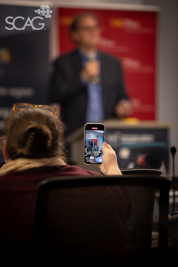 Audience member films speaker on phone in conference setting.