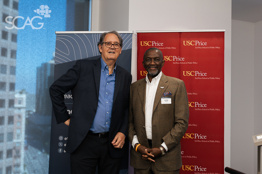 Two men in suits smiling in front of SCAG and USC Price banners.