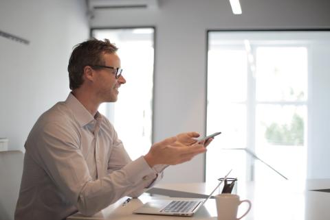 Man in a bright office holding a phone, smiling, with a laptop and mug nearby.